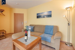 A living room with a sofa, a coffee table with a bottle and glasses, a painting on the wall, and a floor lamp.