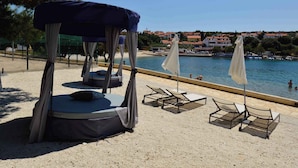 Area sulla spiaggia con lettini e una zona salotto coperta, con vista su un mare calmo e edifici sullo sfondo.