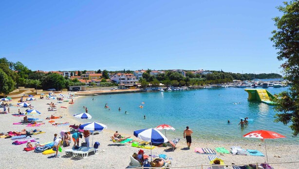 Una spiaggia con persone che prendono il sole e nuotano, ombrelloni e barche sullo sfondo.