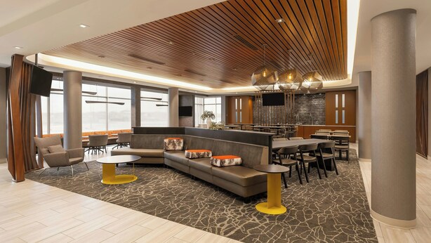 Un hall d’hôtel moderne avec un plafond incurvé, un espace salon avec des canapés et des fauteuils, un coin repas avec des tables et des chaises, et une cuisine en arrière-plan.