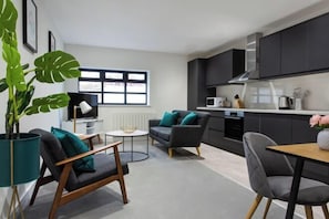 Een moderne woonkamer met een bank, een salontafel en een eettafel. Er staat een grote potplant en een raam met zonwering.