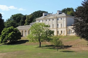 Un grand bâtiment historique, doté de nombreuses fenêtres et d’une façade symétrique, entouré d’une pelouse bien entretenue et d’arbres.