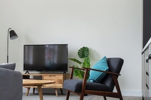 A modern living room with a flat-screen TV on a wooden stand, a dark armchair with a blue pillow, a small wooden table, and a large green plant. - Viridian Apartments in Queen's Park Serviced Apartments (Brent)