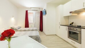 Une pièce de taille modeste comprenant un lit, une petite table ornée d’un vase de fleurs rouges, une kitchenette équipée d’une cuisinière et d’un four, et un téléviseur fixé au mur.