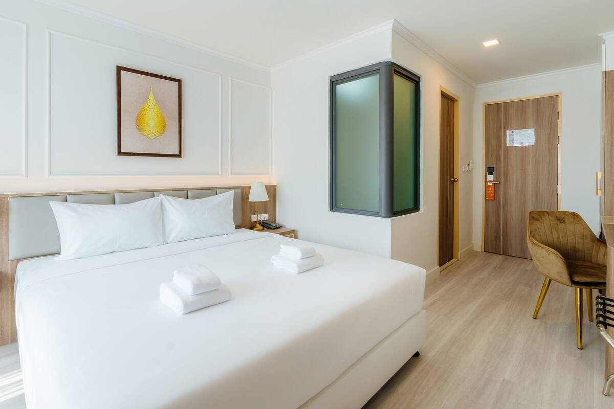 Quarto de hotel com cama grande, mesas de cabeceira com luminárias, escrivaninha com cadeira, quadro na parede e porta com placa.
