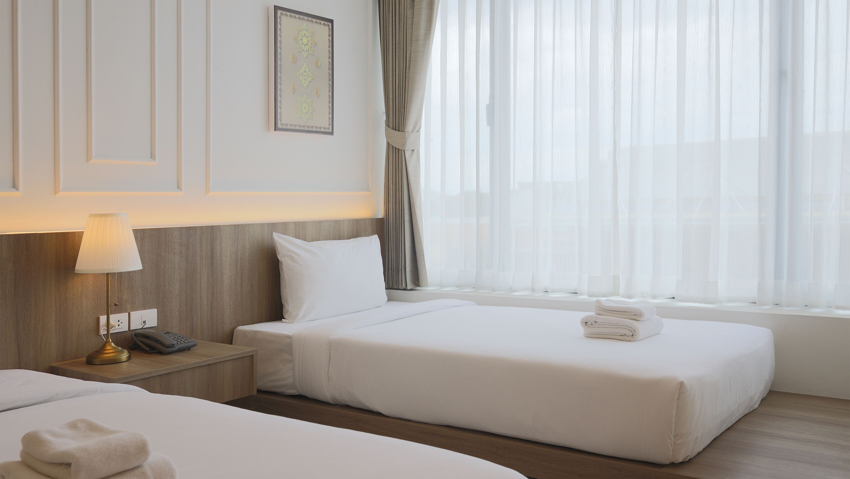 Hotellihuone, jossa on kaksi sänkyä, yöpöytä lampun kanssa, seinällä kehystetty taulu ja ikkuna, jossa on läpikuultavat verhot.