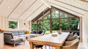 A spacious room with a large wooden table and chairs, a grey sofa, and a window offering a view of greenery. - 5 Star Holiday Home in Vaeggerlose (Vaeggerlose)