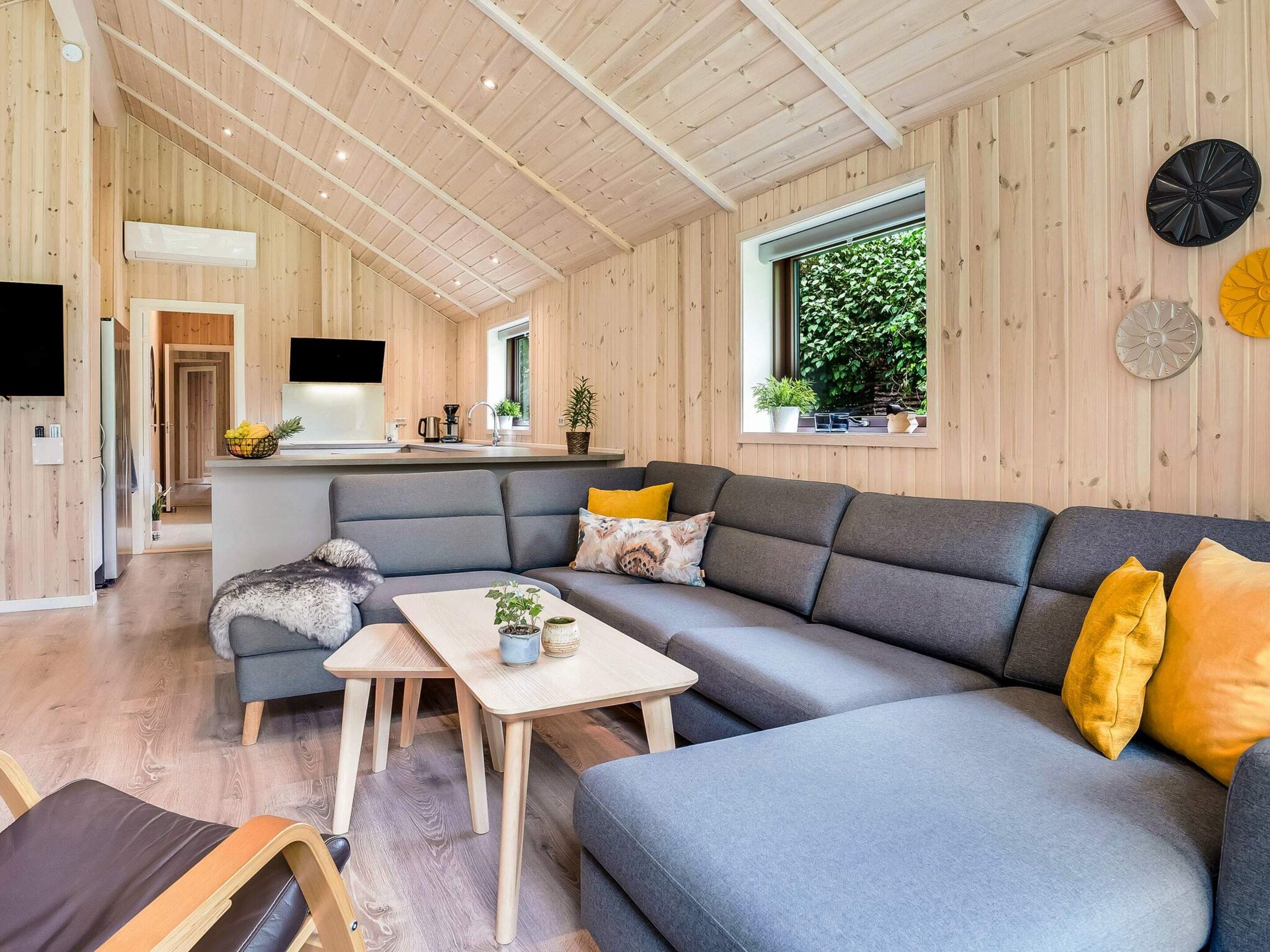 Un intérieur moderne avec un canapé d’angle gris, une table à manger en bois et un coin cuisine équipé d’un téléviseur.