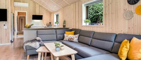 A modern interior with a grey sectional sofa, a wooden dining table, and a kitchen area with a TV.