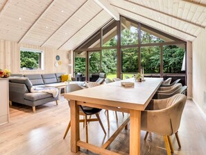 A spacious room with a large wooden table, comfortable seating, and large windows offering a view of greenery. - 5 Star Holiday Home in Vaeggerlose (Vaeggerlose)