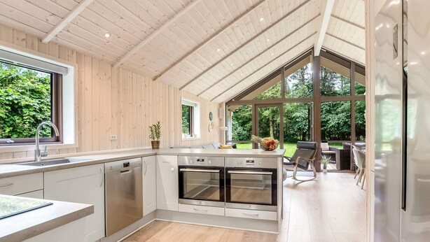 A modern kitchen with wooden paneling, a central island with appliances, and large windows offering views of greenery. - 5 Star Holiday Home in Vaeggerlose (Vaeggerlose)