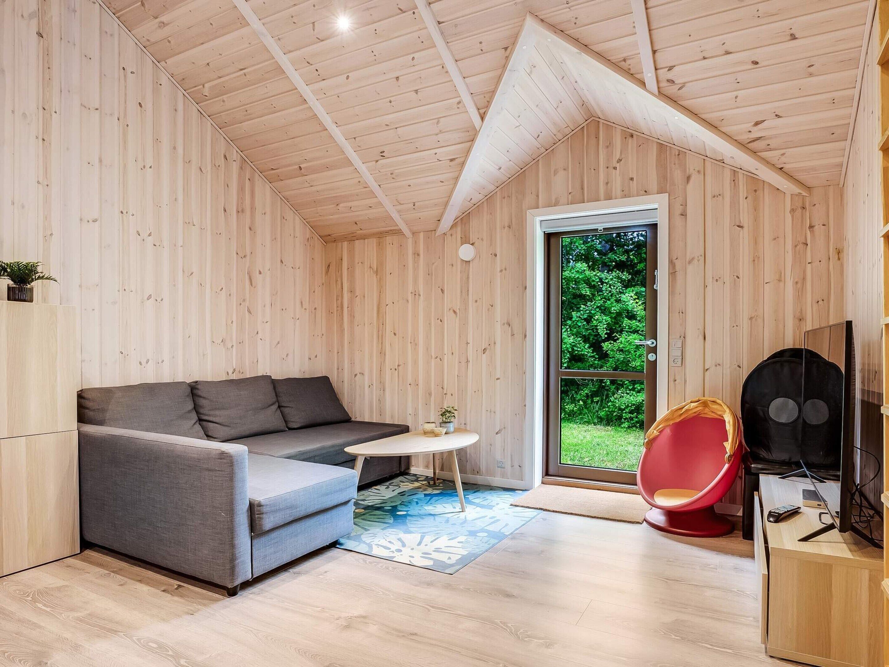 Ein gemütliches Holzambiente mit einem grauen Sofa, einem kleinen runden Tisch, einem roten Sessel und einem Fernseher auf einem Holzschrank.