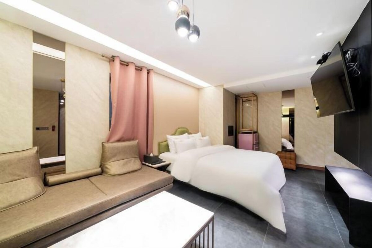 A modern hotel room with a bed, sofa, bedside table, and a TV mounted on the wall.