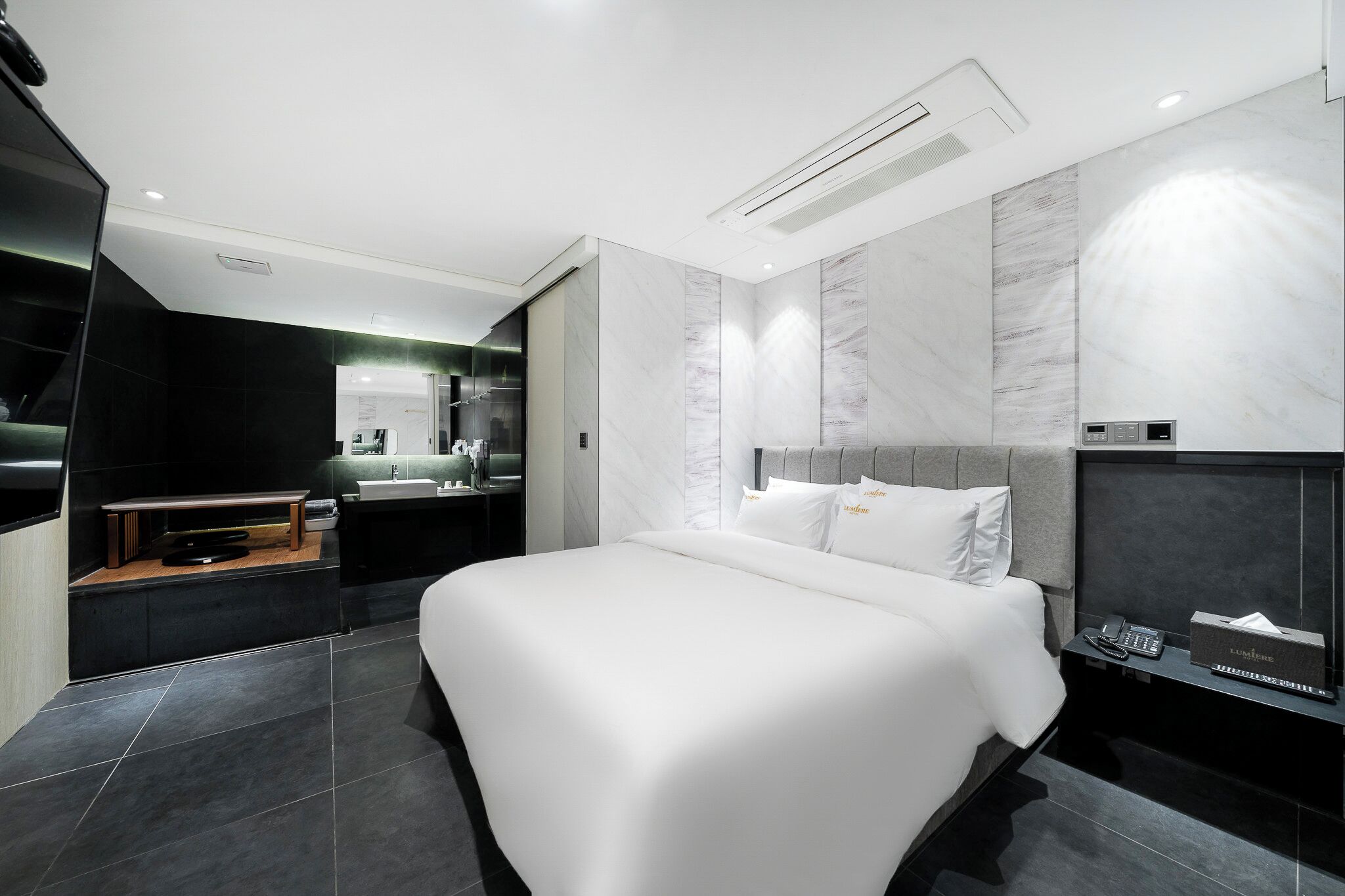 Quarto de hotel moderno com uma cama grande, uma toucador com espelho e uma televisão sobre um suporte de madeira.