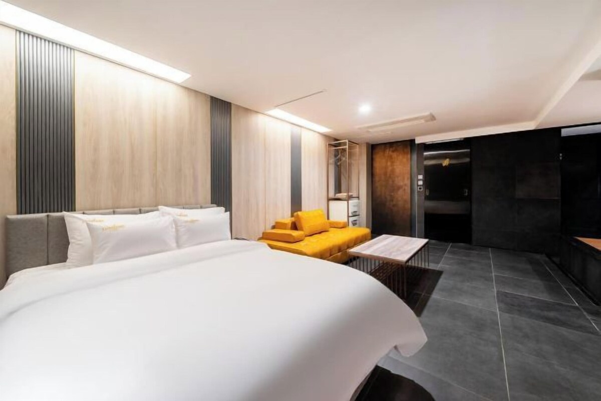 A modern hotel room with a large bed, a sofa, and a small table. There is a shower area and a dark-toned wall.