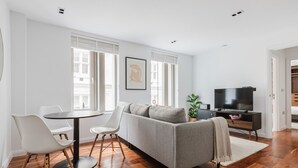 A modern living room with a grey sofa, white chairs, a round table, and a TV on a wooden stand.