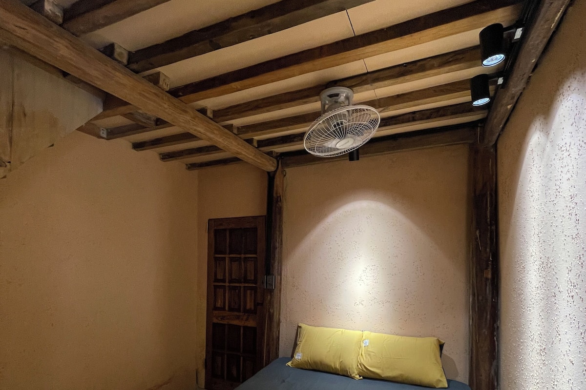 A room with a bed, a wooden ceiling, a fan, and a door.