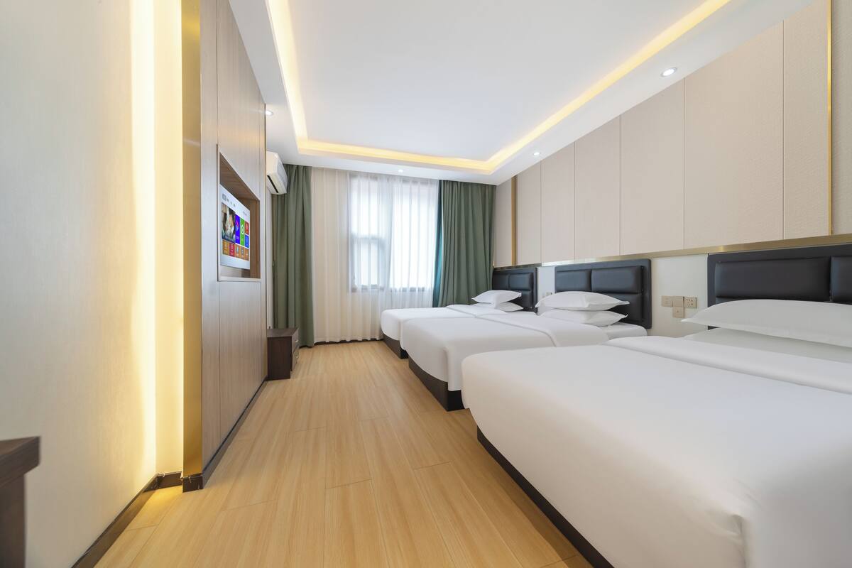 Quarto de hotel com três camas, piso de madeira, uma janela grande com cortinas e televisão presa à parede.