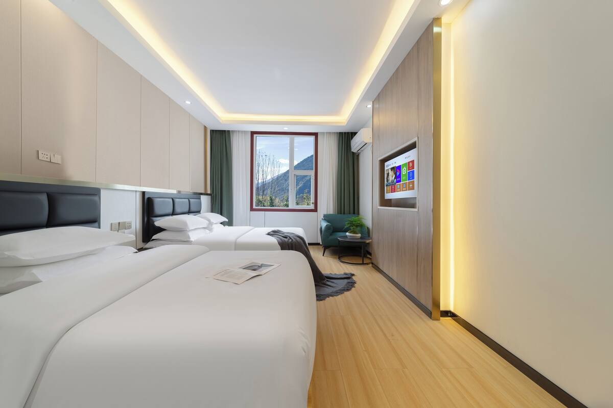 Quarto de hotel com duas camas, televisão, janela com vista para as montanhas e uma pequena mesa com uma planta.