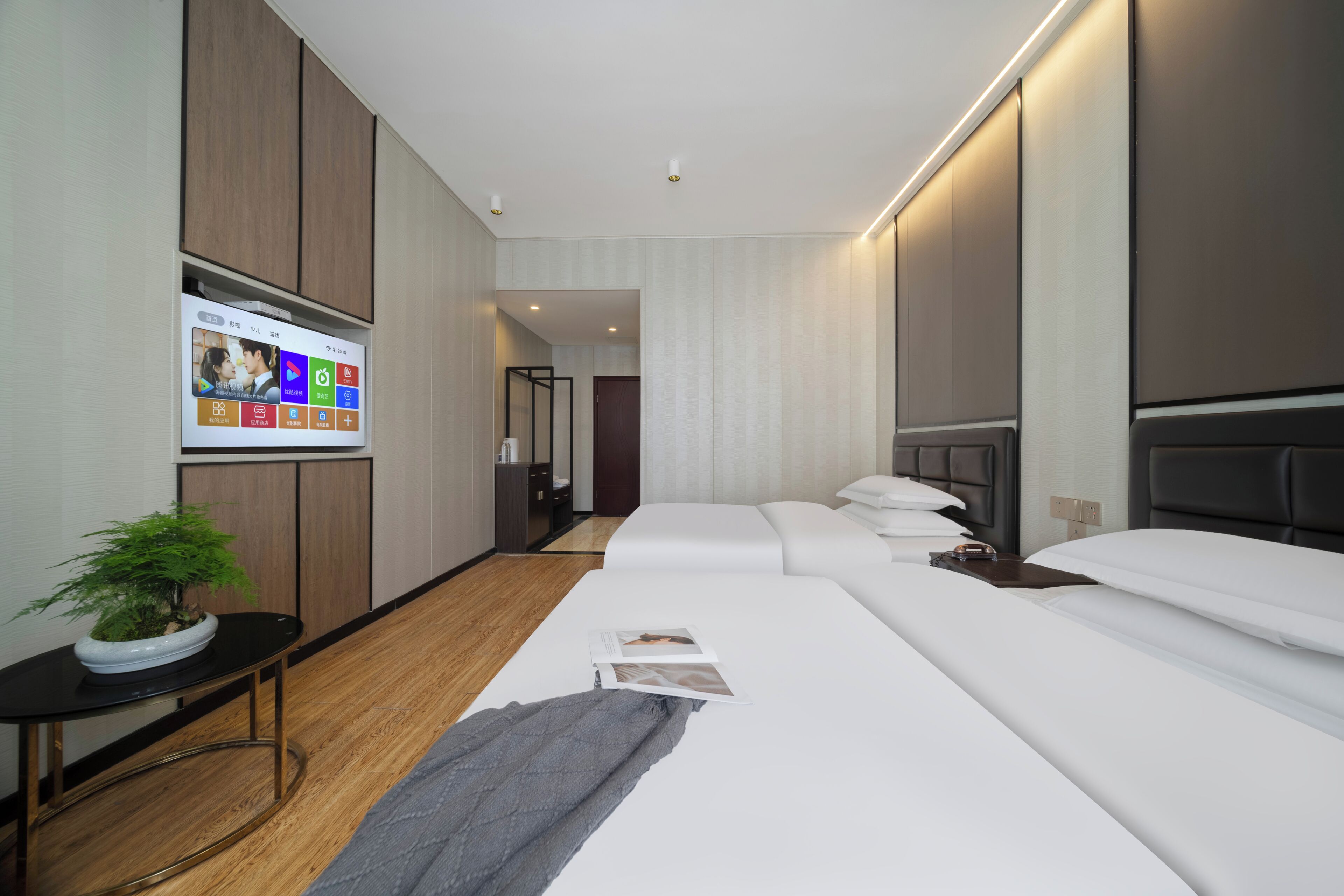 A modern hotel room with a large bed, a bedside table, a TV mounted on the wall, and a potted plant.