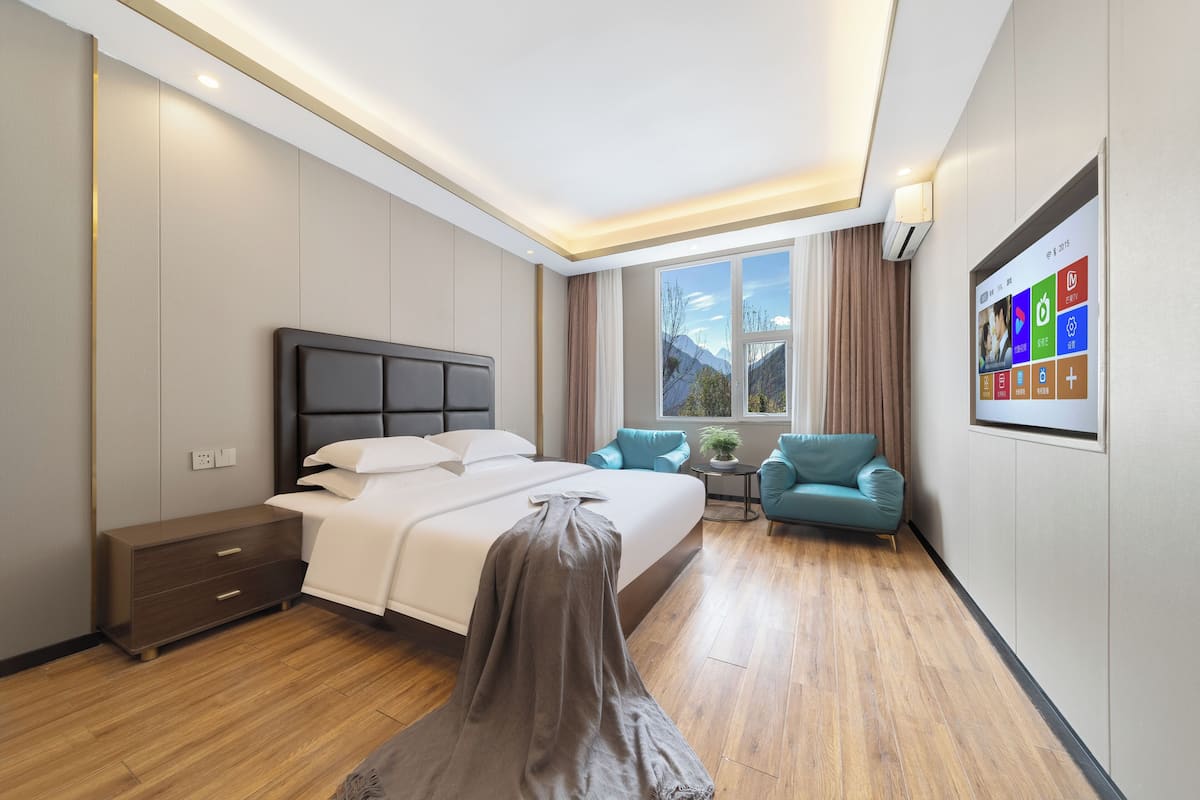 Quarto de hotel moderno com cama, mesa de cabeceira, pequena mesa com planta, duas poltronas e uma grande janela com vista para as montanhas.