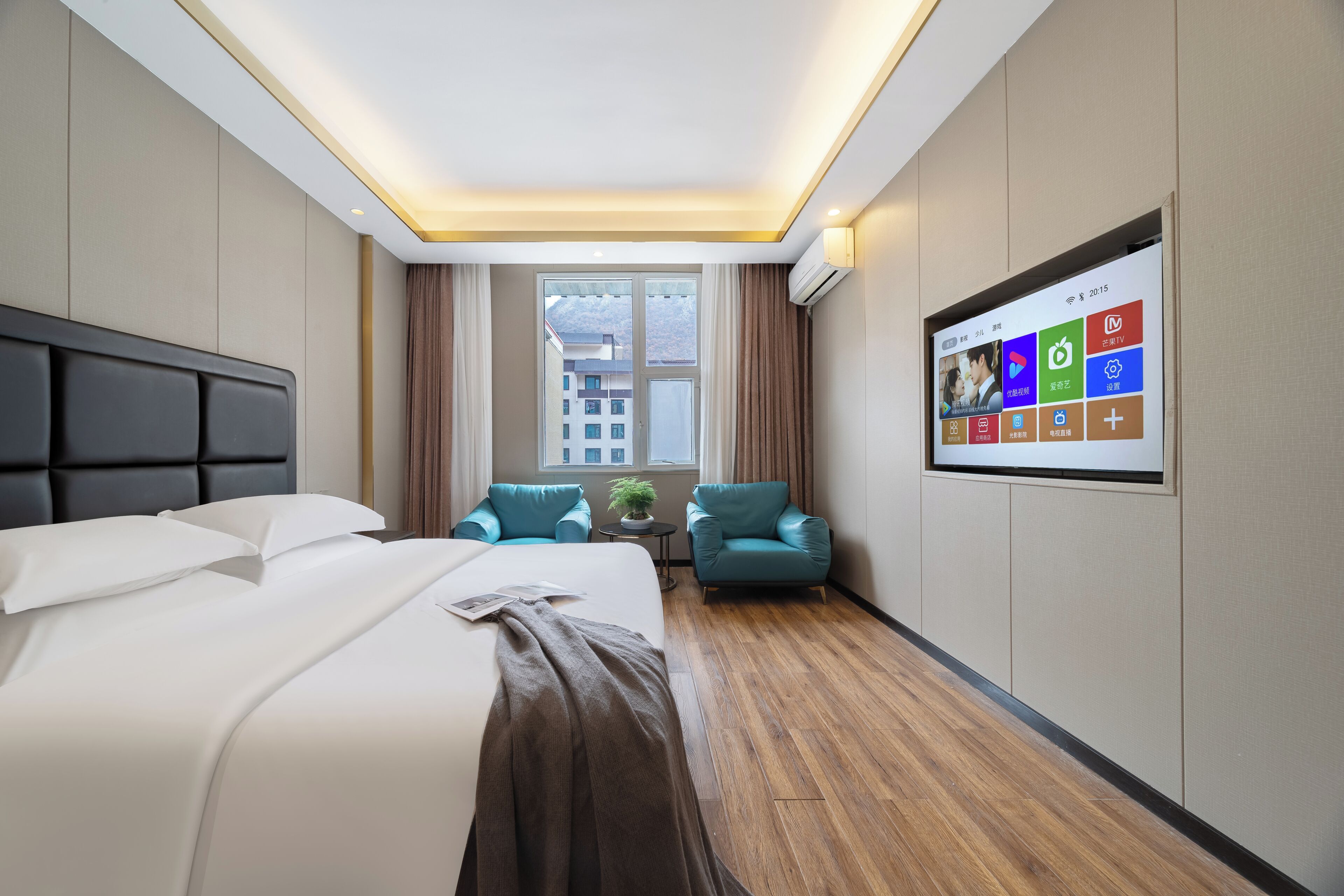 A modern hotel room with a large bed, two armchairs, a small table, a TV, and a window with a city view.