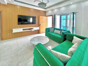 A modern living room with a flat-screen TV, green sofas, a coffee table, and a wooden panel wall.