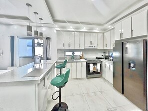 Une cuisine moderne avec des meubles blancs, un réfrigérateur en acier inoxydable, un four à micro-ondes et une cuisinière à gaz. On y trouve trois tabourets de bar verts et une fenêtre avec des rideaux.