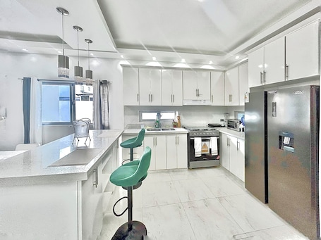 A modern kitchen with white cabinets, a stainless steel refrigerator, a microwave, and a gas stove. There are three green bar stools and a window with curtains.. Beach 100 mts away! Live The Luxury In Rio San Juan Parking Included!