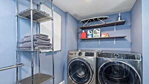 A laundry area with two front-loading washing machines, a metal shelf with folded towels, and shelves with laundry supplies.