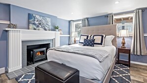 A bedroom with a fireplace, a large bed, a nightstand, and a view of the outdoors.