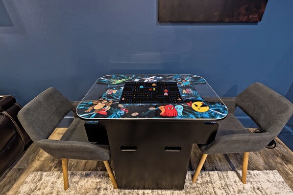 Une table avec un plateau aux couleurs vives et décoré sur le thème des jeux, deux chaises grises et un meuble noir doté de poignées.