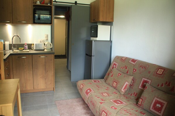 A compact kitchen with wooden cabinets, a microwave, and a refrigerator. A sofa with patterned cushions is in the living area. - Gentianes Gnb66 Cosy & Mountain 3 Pers (Chatel)