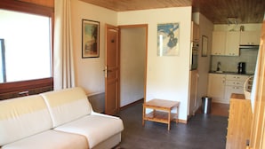 A living room with a white sofa, a small wooden table, and a coffee-themed rug. There is a kitchen area with cabinets and a sink visible. - Rhododendrons Rd501 Center & Telecabine 4 Pers (Chatel)
