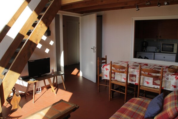 Une pièce chaleureuse avec un plafond en bois, un téléviseur posé sur un meuble en bois, une table à manger avec des chaises et un coin cuisine équipé d’un four à micro-ondes.