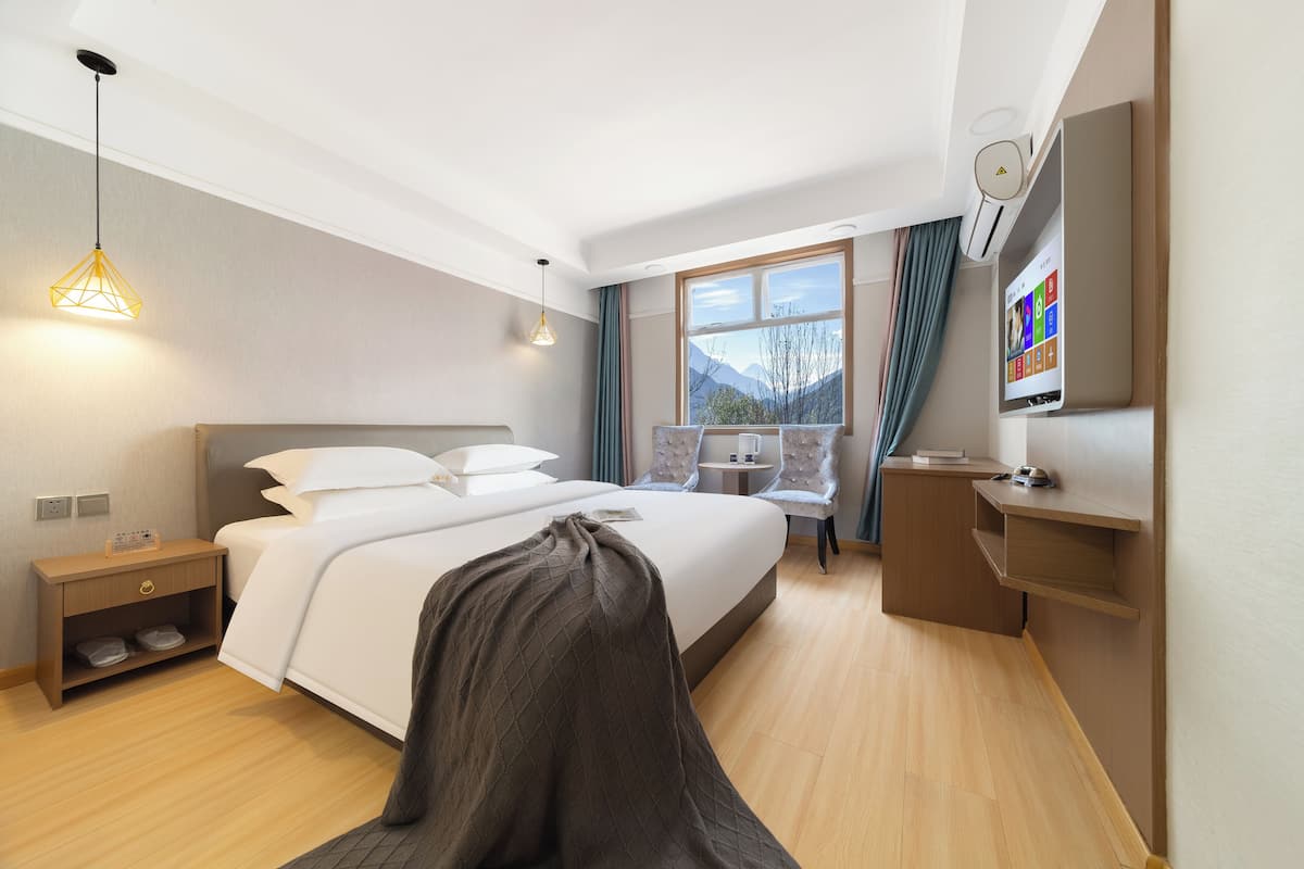 Ein Hotelzimmer mit Bett, Nachttisch, Lampe, Fernseher und einem Fenster mit Bergblick.