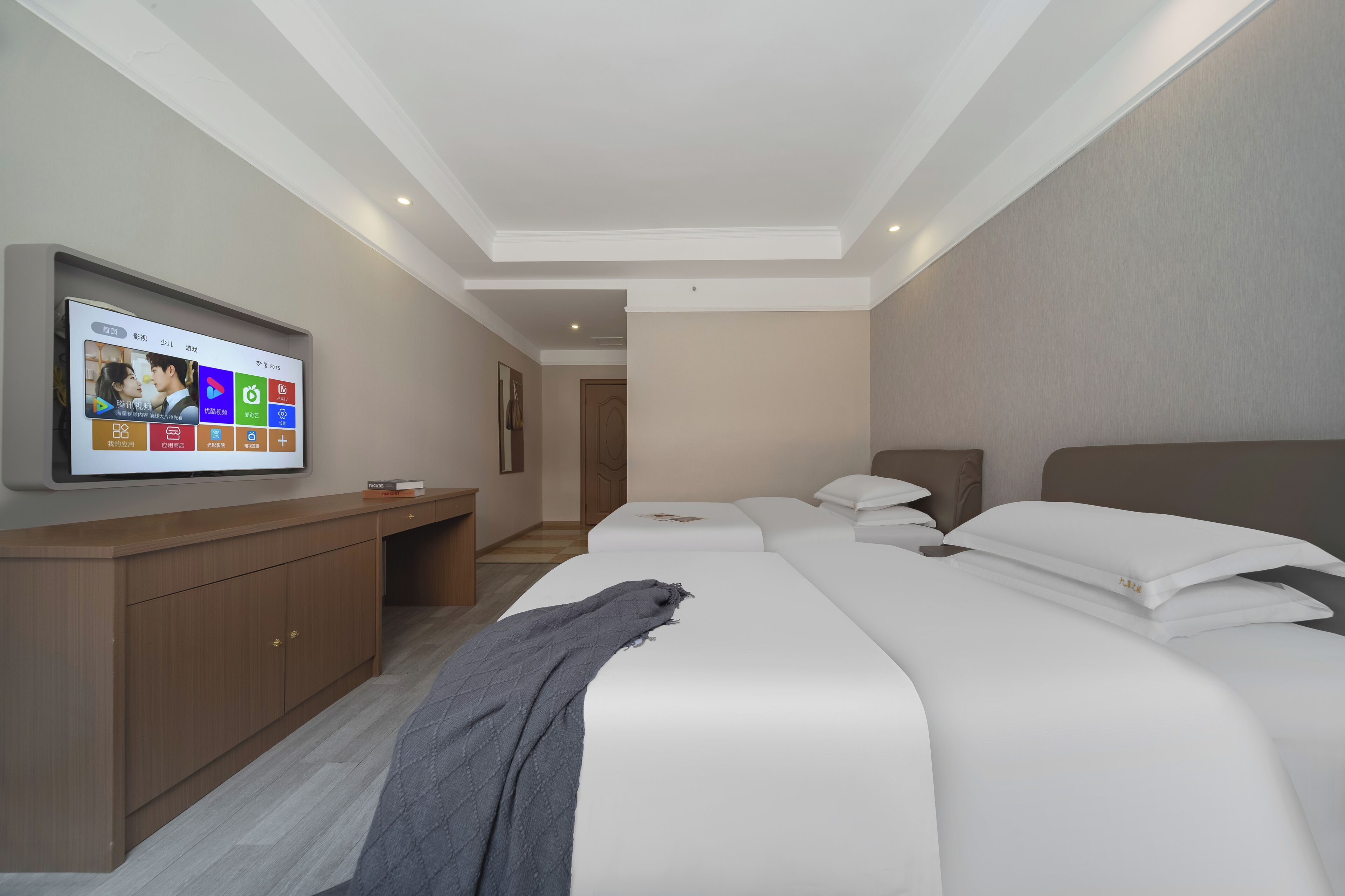 Habitación de hotel con televisión instalada en la pared, un escritorio de madera, una cama con ropa de cama blanca y una manta gris en el suelo.