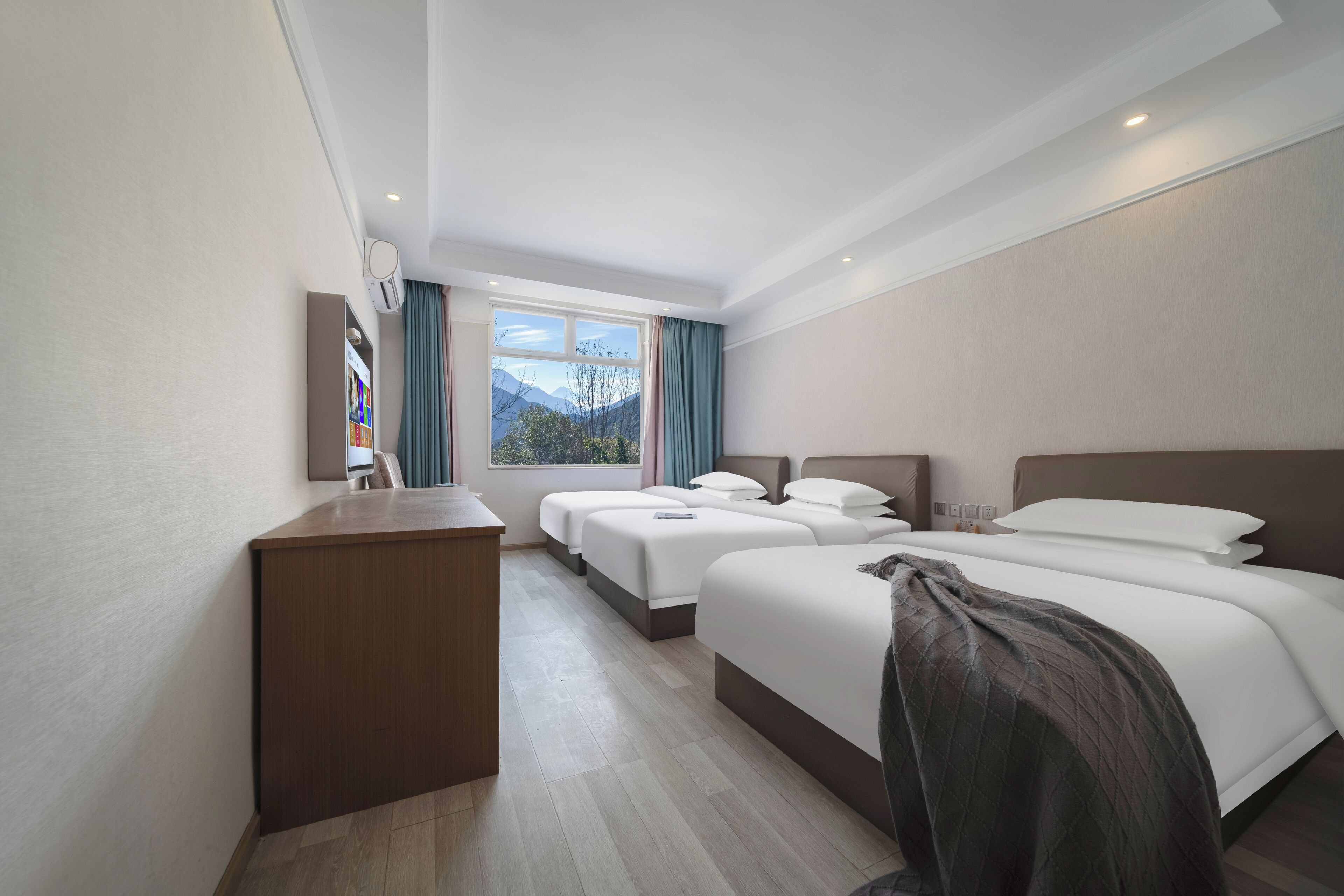 A hotel room with a desk, two beds, a view of mountains, and a window with curtains.