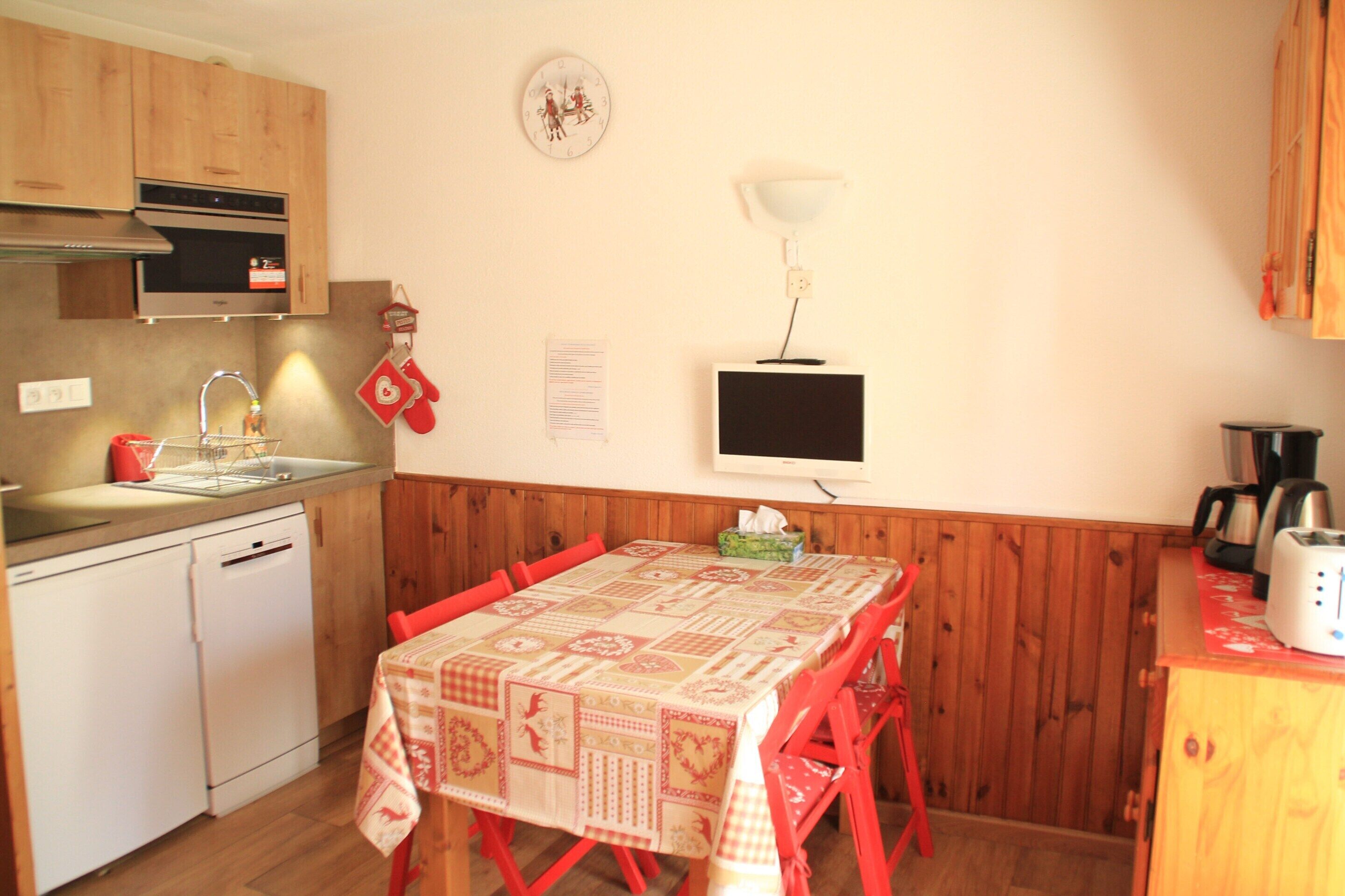 Een kleine keuken met houten kastjes, een inbouwoven, een spoelbak en een eettafel met rode stoelen.
