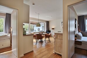 Quarto de hotel moderno com piso de madeira, área de refeições com mesa e cadeiras, televisão e uma grande janela com cortinas.