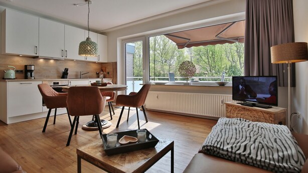 Un salon moderne avec un canapé, une table basse et un téléviseur. Il y a également un coin repas avec des chaises et une table. La pièce est dotée de grandes fenêtres offrant une vue sur les arbres.