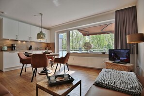 A modern living room with a sofa, a coffee table, and a TV. There is a dining area with chairs and a table. The room has large windows with a view of trees. - The Island Seaside Apartment 7 - The Island (Timmendorfer Strand)