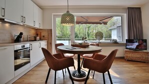 Une cuisine moderne avec des meubles blancs, un coin repas comprenant une table ronde et quatre chaises, un luminaire suspendu et un téléviseur posé sur un meuble en bois.