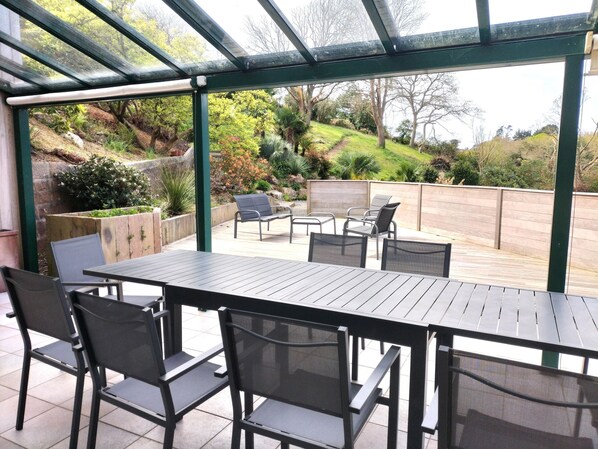 A covered patio with a table and chairs, a wooden deck, and a view of a landscaped garden. - High end Holiday Home for 6 People (Trébeurden)