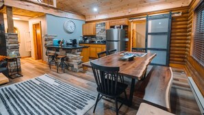 A cozy cabin interior with a wooden dining table, chairs, a kitchen with a stone wall, and a clock on the wall. - Log Haus- Cabin in Amish Country (Danville)