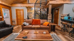 A cozy living room with a brown leather sofa, wooden coffee table, and a spiral staircase. - Log Haus- Cabin in Amish Country (Danville)