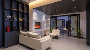 Una sala de estar moderna con chimenea, un sofá y una mesa de centro. Hay estanterías con objetos decorativos y una puerta de cristal que da al exterior.