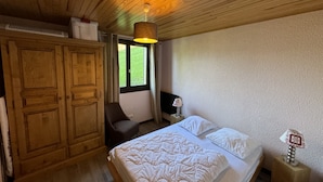 A bedroom with a wooden ceiling, a bed with white bedding, a wooden wardrobe, a chair, a desk with a lamp, and a window with a view of greenery. - Hameau DES 4 Saisons Hsc113 Mountain & View 6 Pers (Chatel)
