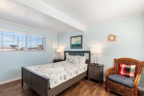 A bedroom with a bed, two bedside tables, a chair, a window with blinds, a painting, and a wall decoration.