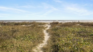 Ein sandiger Pfad führt zu einem Strand mit wenig Vegetation und einem klaren Himmel.